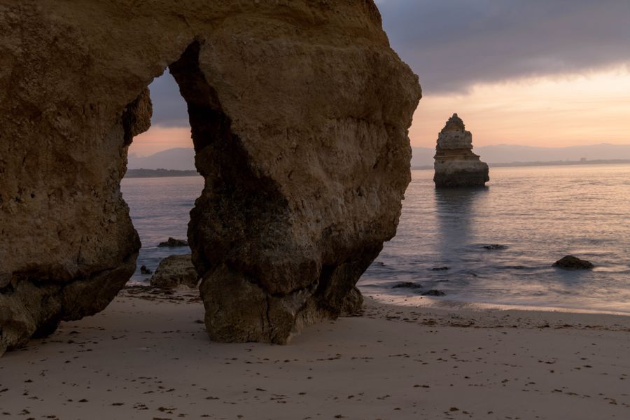 A sandstone arch on a beach frames a lone sea stack standing in calm water under a soft dawn sky