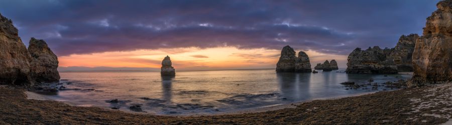 Wide panorama of a curving beach and scattered sea stacks under a band of orange dawn light and dark clouds