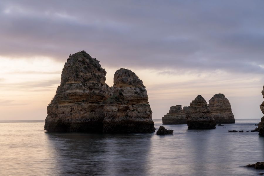 Layered sandstone sea stacks rising from still, silvery water under a pale overcast sky
