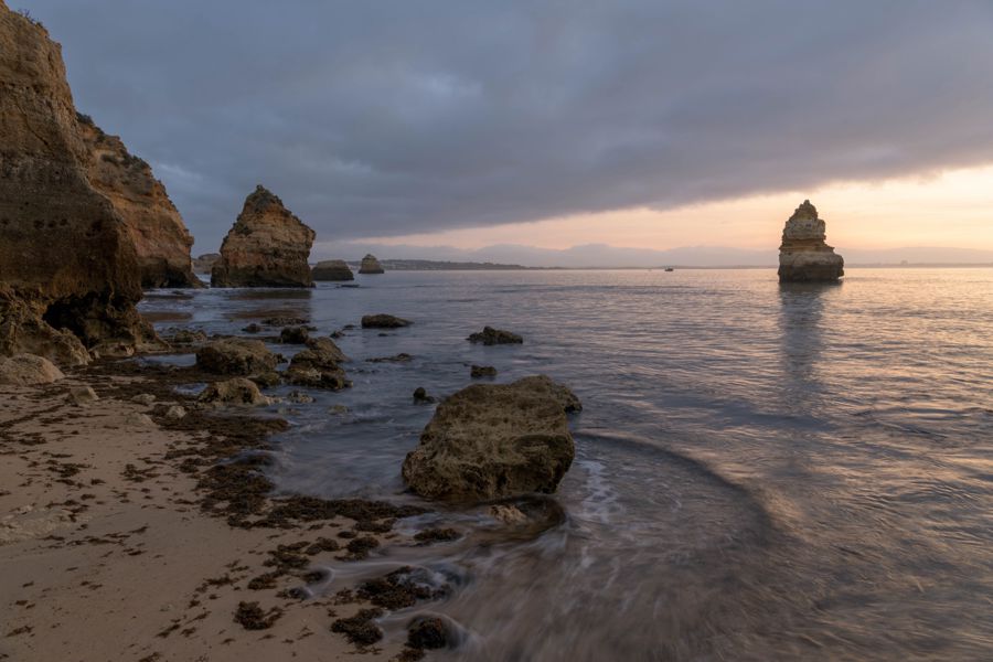 Long exposure of water swirling around rocks on a beach, with sea stacks along the shore and a lone stack offshore