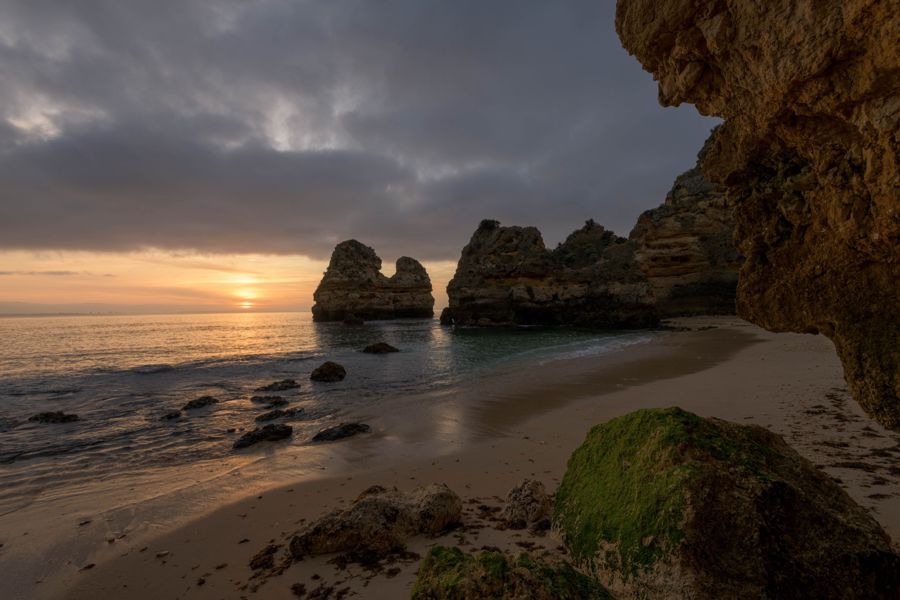 The sun rising between sea stacks, seen from beneath an overhanging cave wall with a mossy boulder in the foreground