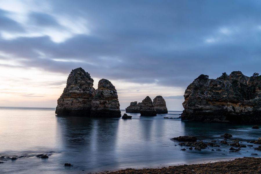 Long exposure of dark sea stacks reflected in smooth blue water before sunrise, under streaked clouds