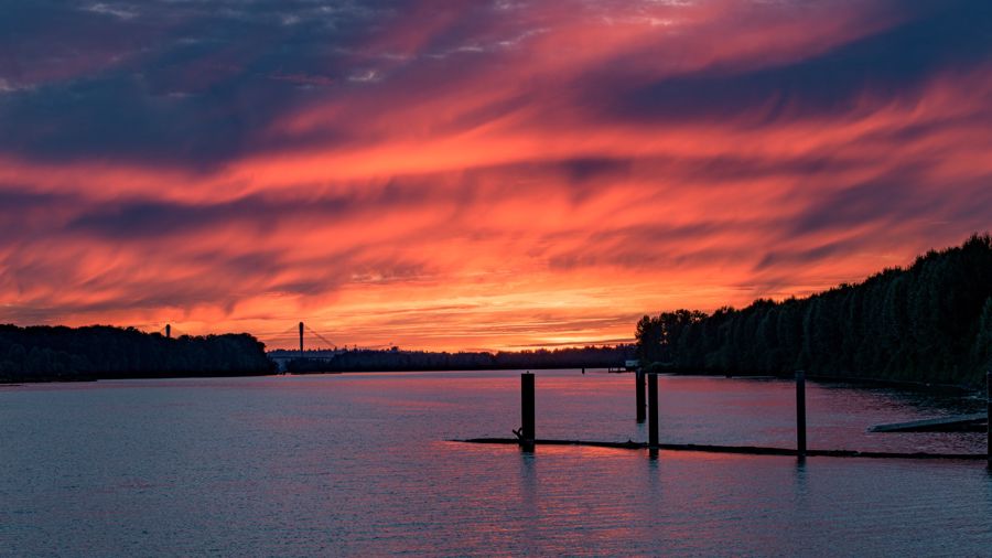 A blazing red and orange sunset sky over a wide river with old pilings, a bridge on the far horizon
