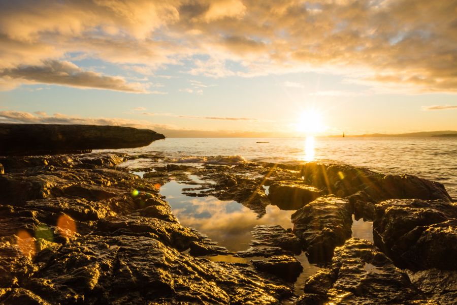 The sun low over the sea as a starburst, lighting up a rocky shoreline and a tide pool reflecting the clouds
