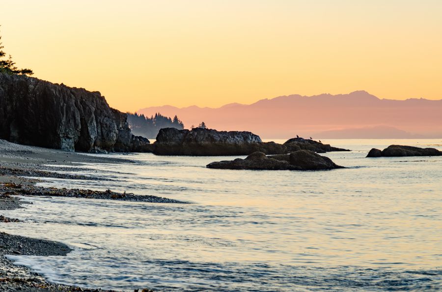 A rocky point and dark cliffs along a pebble beach at dusk, with hazy pink mountains across the water