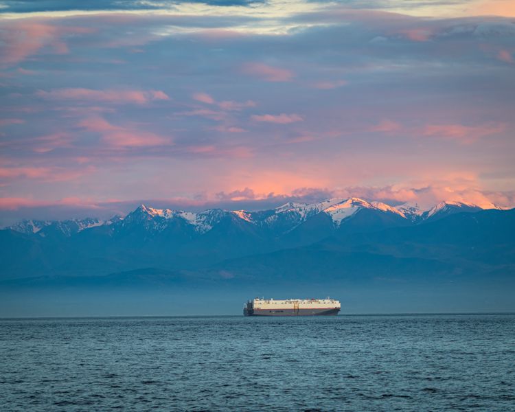 A car carrier ship crossing open water below a range of snow-capped mountains under pink clouds