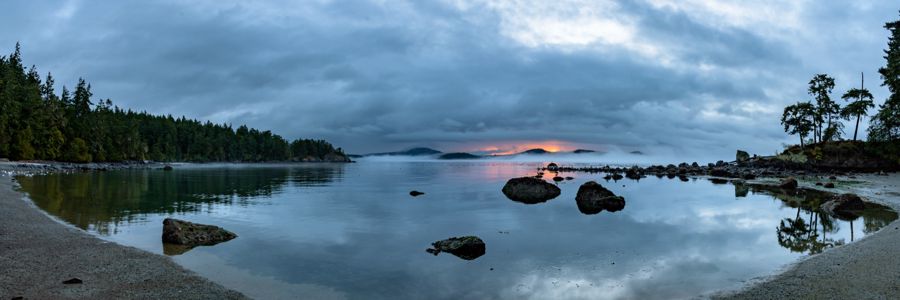 Panorama of a calm cove with scattered rocks, forested shores and distant islands reflected under grey-blue clouds