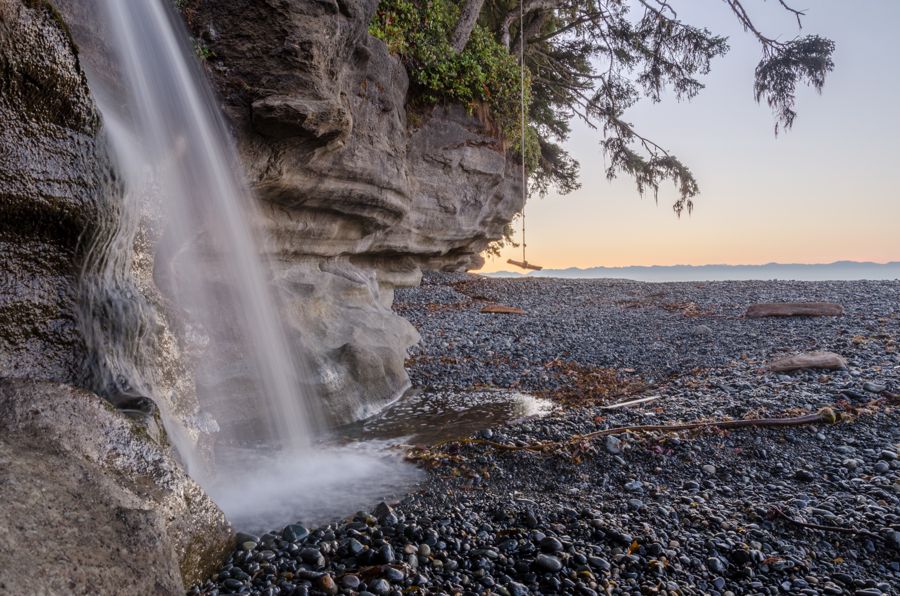 A waterfall pouring over a sandstone overhang onto a pebble beach, with a rope swing and the sea glowing at dusk