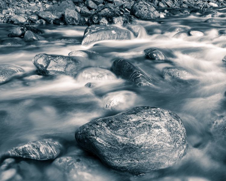 Blue-toned, nearly black and white long exposure of a rocky creek, the water smoothed into mist around round boulders